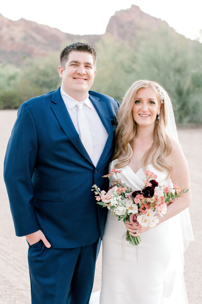 Bride and groom with arms around each other standing in desert landscape. Bride holding a bouquet.