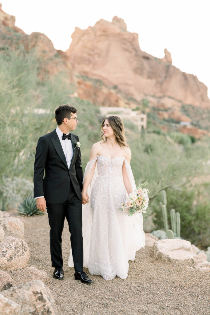 Bride and groom holding hands, bride holding bouquet in other hand, while walking on desert mountain path of pebbles.