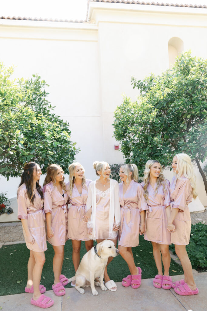 Bride with bridesmaids standing in line in bathrobes, and white dog sitting in front of them.