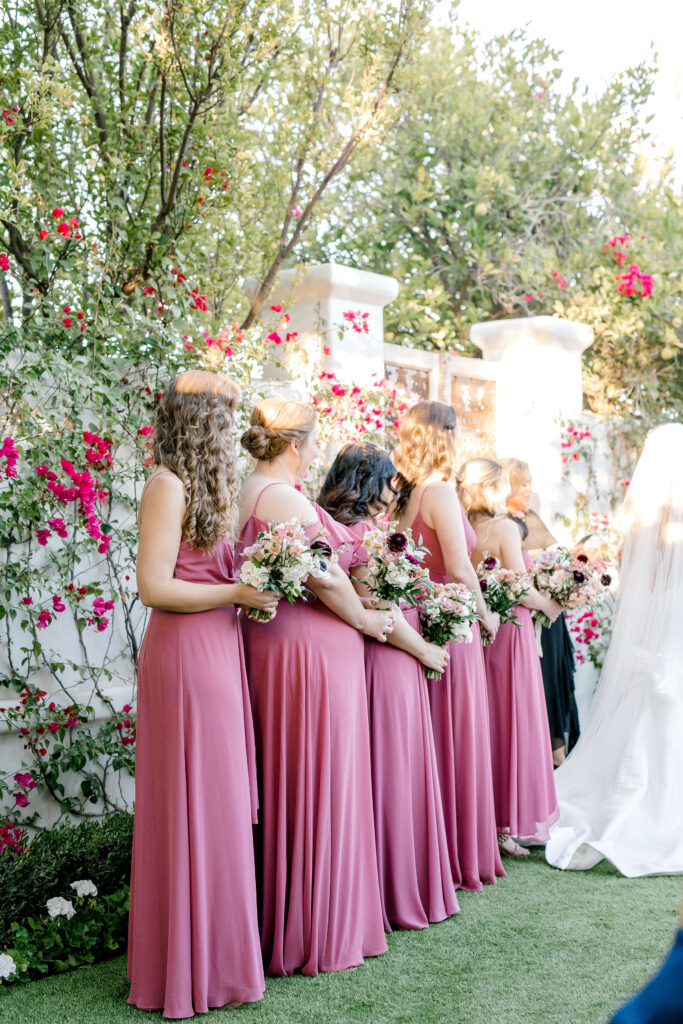 Bridesmaids standing in a row at front of ceremony holding bouquets wearing berry pink colored dresses.