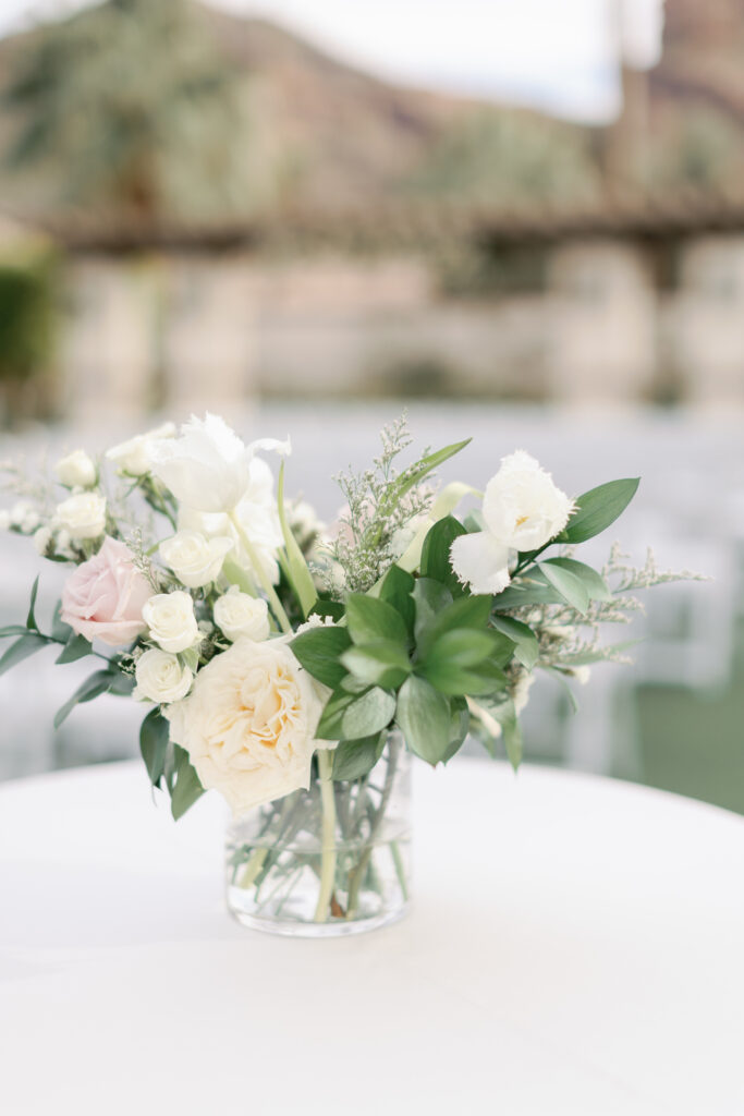 Floral arrangement in glass vase on table with white linen of white and blush flowers and greenery.
