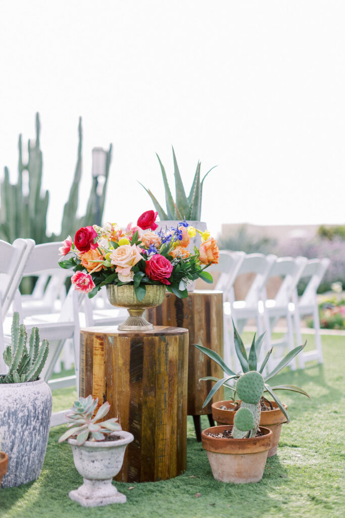 Back of wedding ceremony aisle decor including floral arrangement in gold vase elevated on wood pillar and another desert plant on wood pillar. Various cacti on ground in terracotta and cement pots.