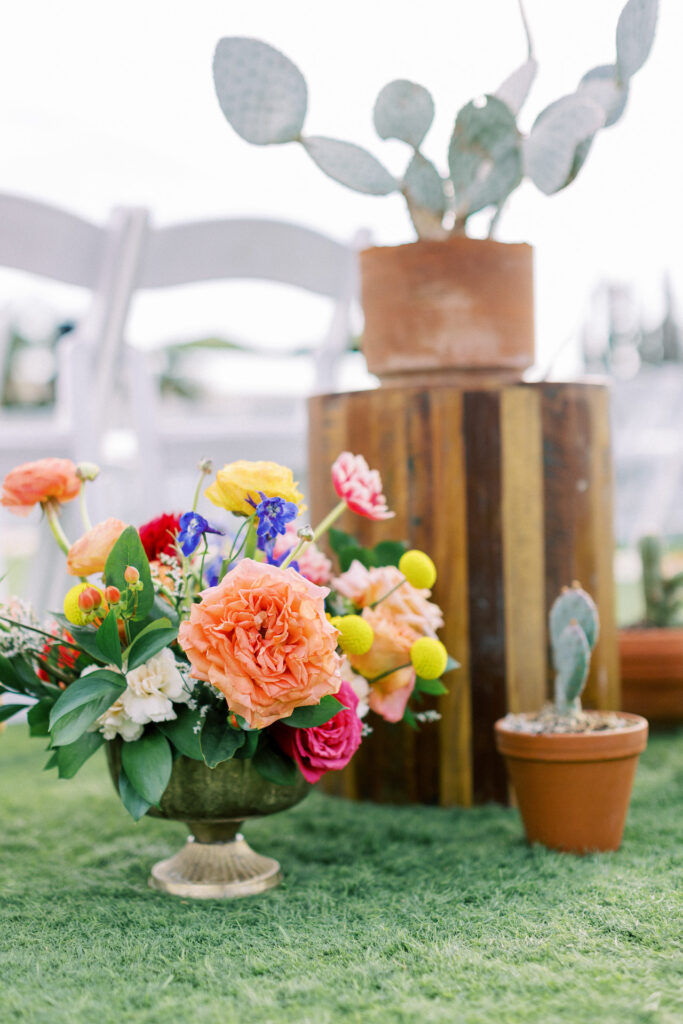 Back of wedding ceremony aisle decor including floral arrangement in gold vase on ground, elevated on wood pillar cactus and cactus on ground in terracotta pots.