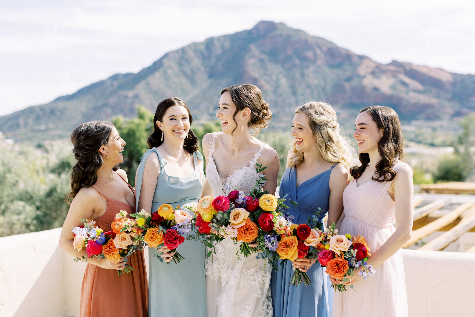 Bride standing outdoors with bridesmaids, all smiling and holding bouquets.