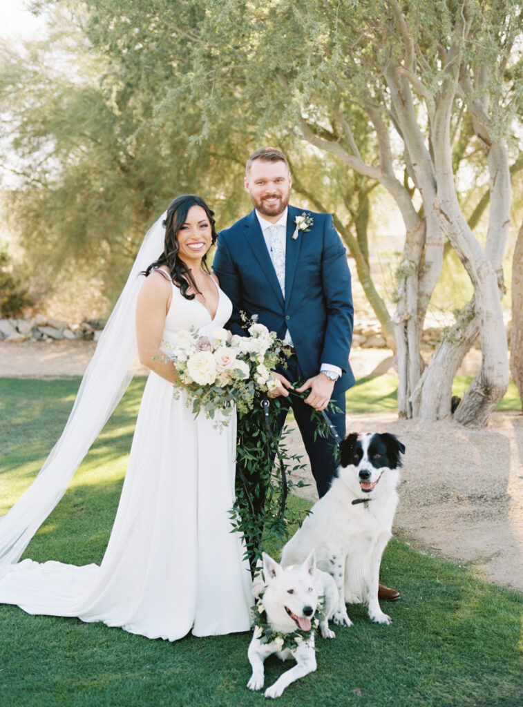 Bride and groom standing in grass with desert landscape behind them, smiling while holding two dogs on leashes and bride holding a bouquet.