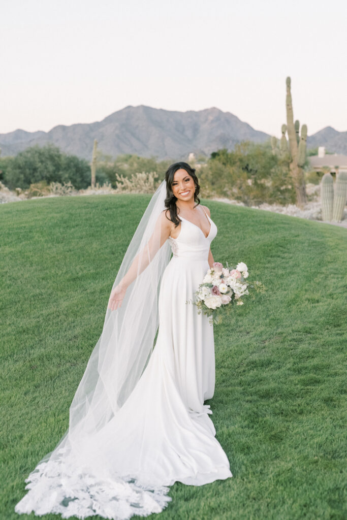 Bride standing on grass hill, holding veil out with arm and bouquet in other hand, smiling.