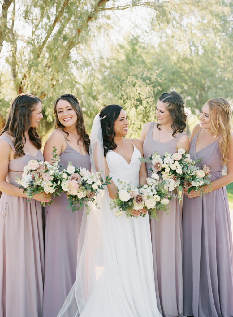 Bride standing in a line with bridesmaids wearing light purple, all smiling and holding bouquets.