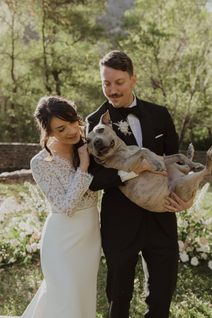 Bride and groom standing next to each other, smiling and looking down at dog groom is holding.