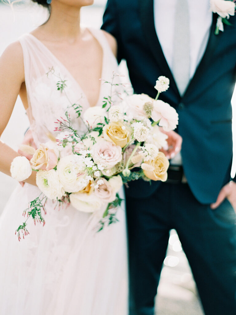 Bride and groom in background with bridal bouquet of white, yellow, and blush floral in foreground.