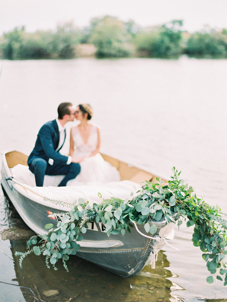 Bride and groom sitting in row boat kissing with greenery added to the front of it.