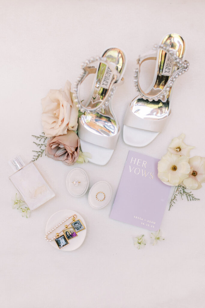 Bridal flat lay including shoes, perfume, vows, rings, and charms.