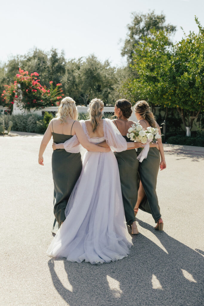 Bride walking with arms around bridesmaids in olive green dresses.
