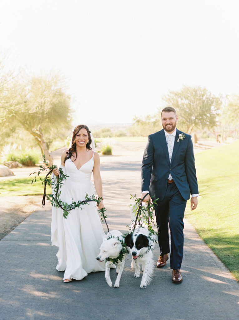Bride and groom walking with two dogs on leashes, both leashes covered in greenery.