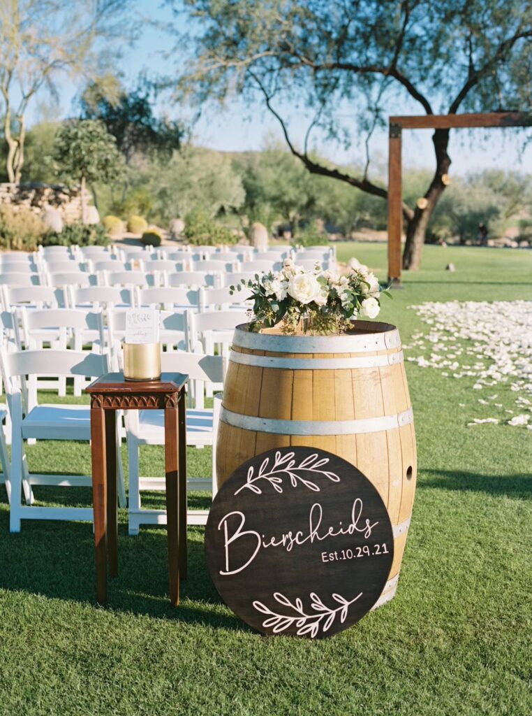 Back of outdoor ceremony space at McDowell Mountain with floral arrangement placed on wood barrel and custom sign leaned up against it.