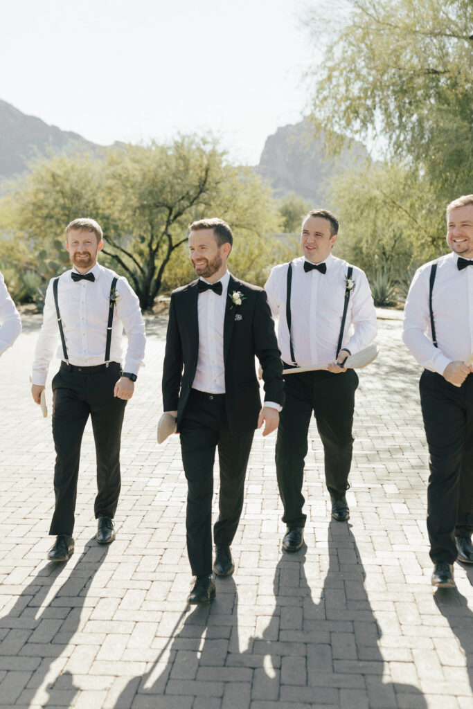 Groom in black suit walking with groomsmen wearing white shirts, black pants, suspenders, and bow ties.