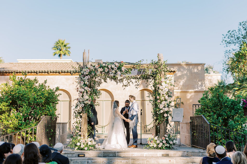 Bride and groom under chuppah with floral installed on it at ceremony in altar space.