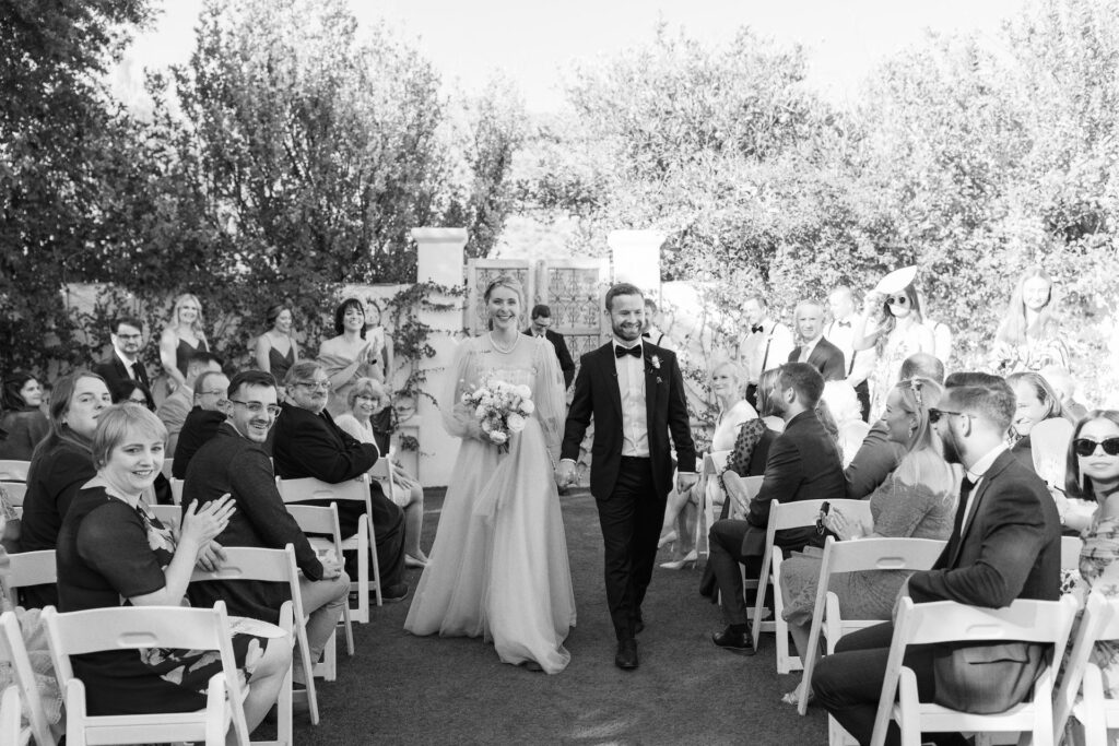 Bride and groom walking down outdoor wedding ceremony aisle holding hands, smiling at guests sitting in chairs.