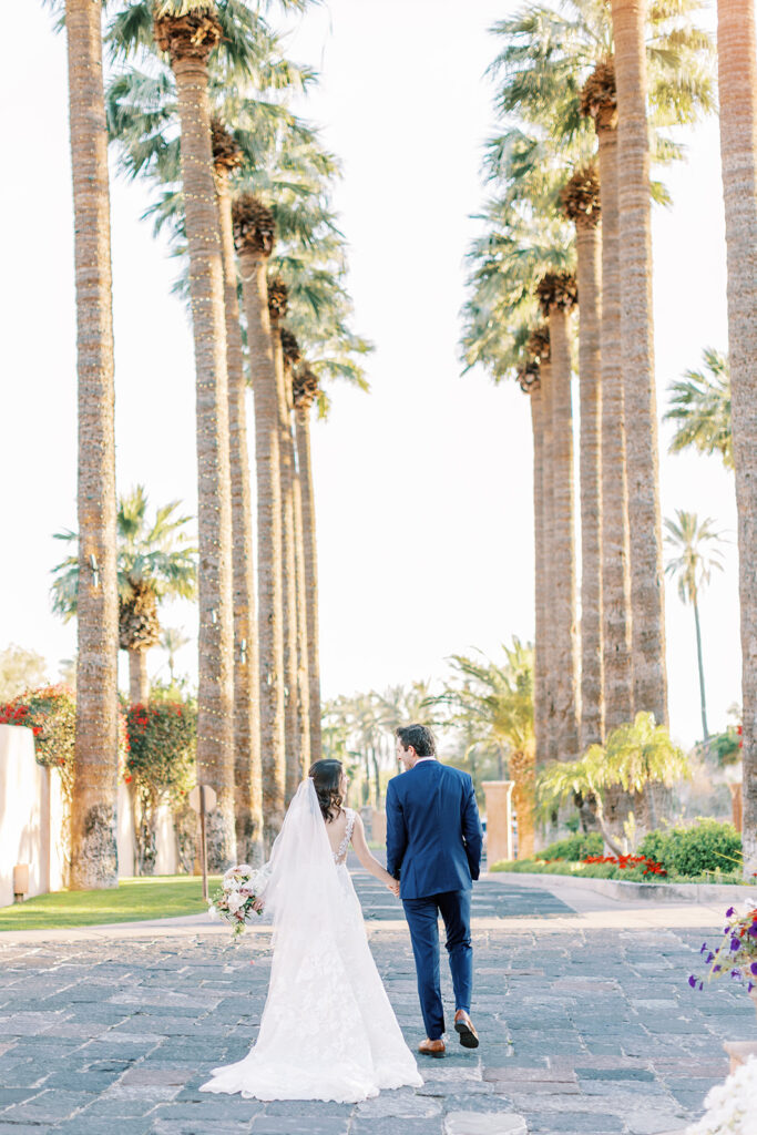 Bride and groom holding hands walking down stone path between tall palm trees.