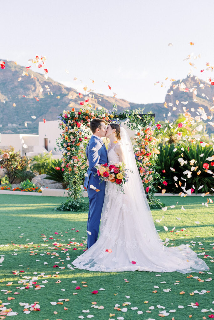 Bride and groom kissing in front of wedding arch decorated with flowers with rose petals falling around them.