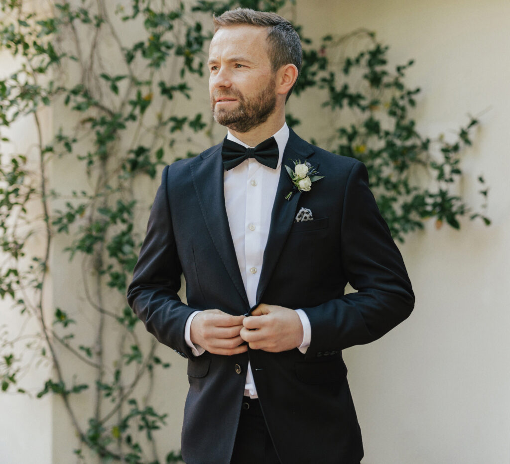 Groom buttoning black suit jacket with bow tie, looking off to side.