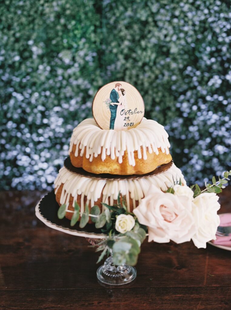 Stacked bunt cakes with custom wedding topper in front of greenery wall.