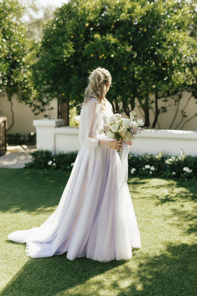 Bride looking behind herself, holding white flowers bouquet, in custom lavender wedding gown.