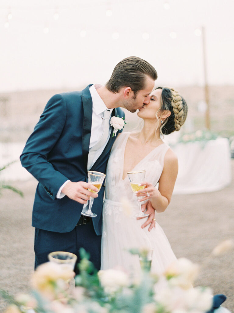 Bride and groom kissing while holding wine glasses at outdoor reception.