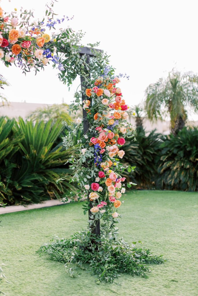 Square wedding arch with floral installation of greenery with vibrant colors including fuchsia, yellow, peach, purple, and blue.