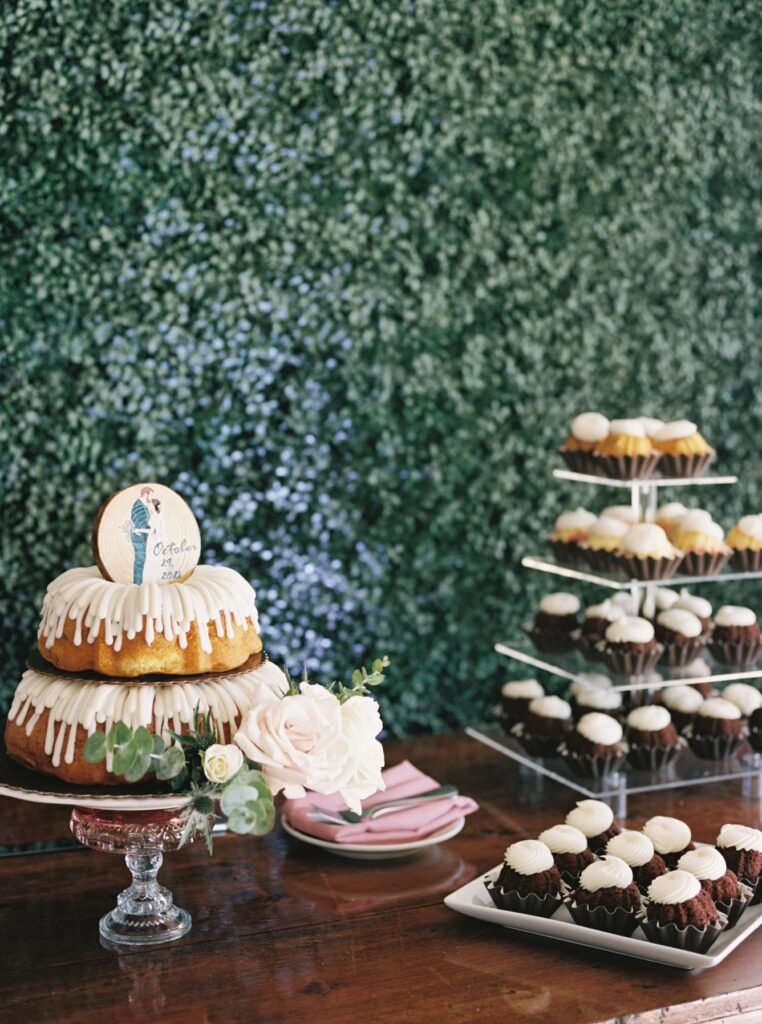 Wedding dessert table of cupcakes and stacked bunt cakes in front of greenery wall.