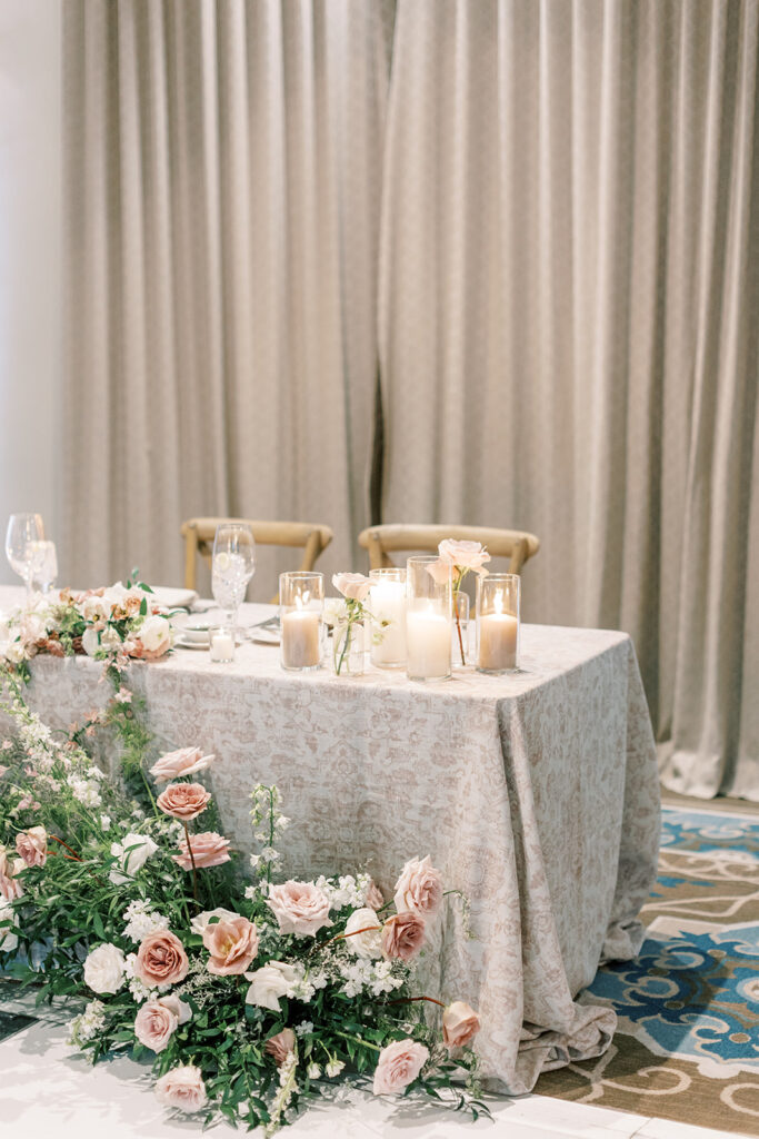 Sweetheart table with tape and cream tablecloth and candles and bud vases on top and ground floral arrangements in front.