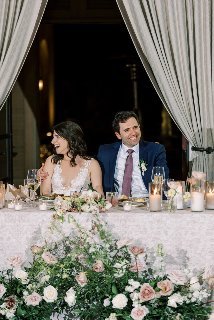 Bride and groom sitting at reception sweetheart table with candles and bud vases, smiling to side.