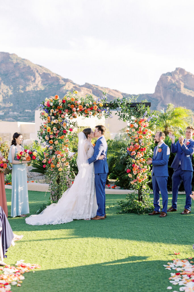 Bride and groom kissing under wedding arch during outdoor ceremony with mountains in distance.