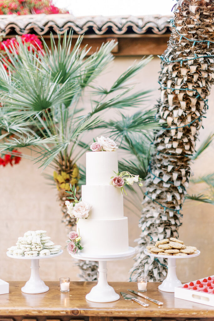 Three tiered white wedding cake with pink and white floral added on outdoor dessert table.