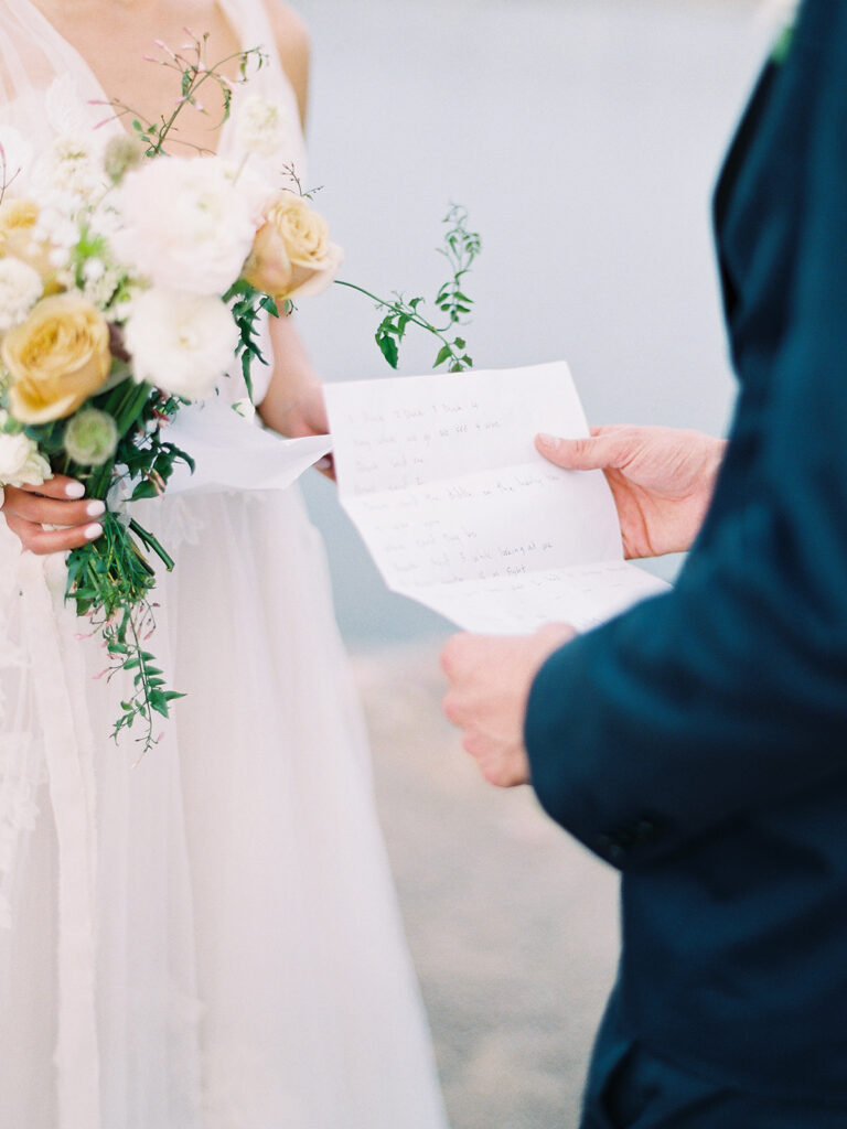 Groom holding paper with wedding vows written on it with bride standing across from him holding bouquet.