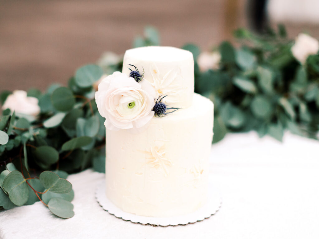 Two tiered white wedding cake with white flower and blue thistle added.