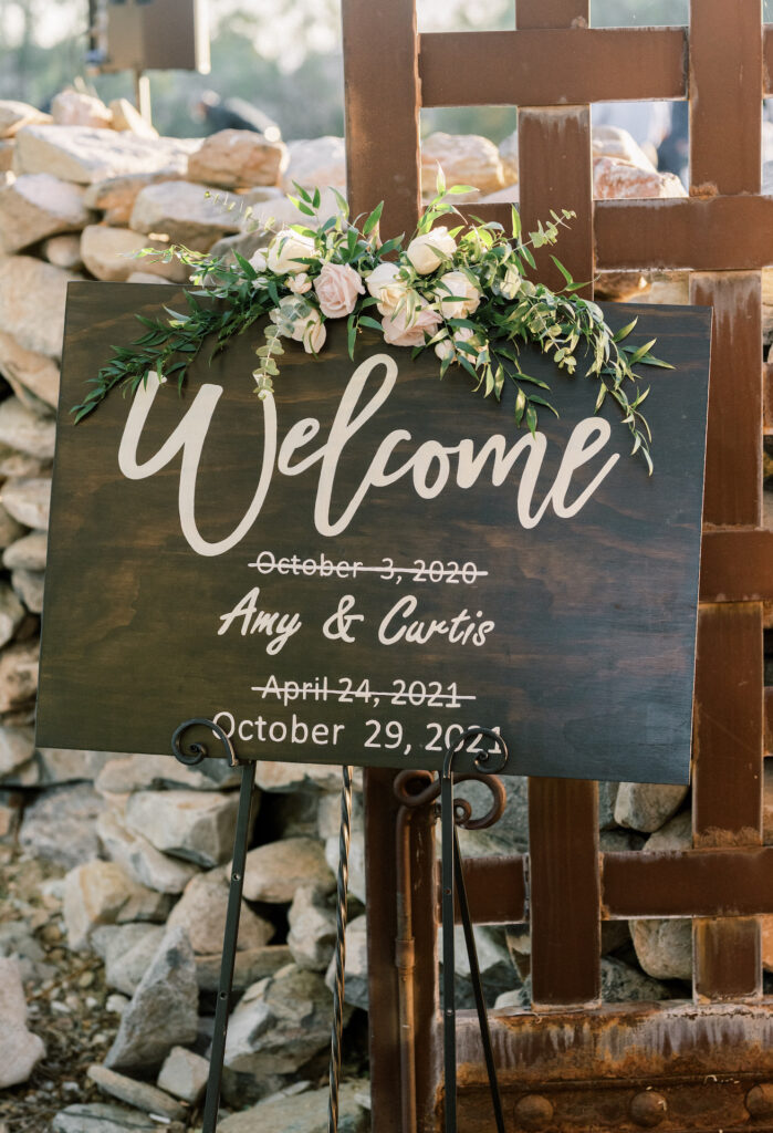 Custom wood rectangle welcome sign with floral and greenery attached to top.