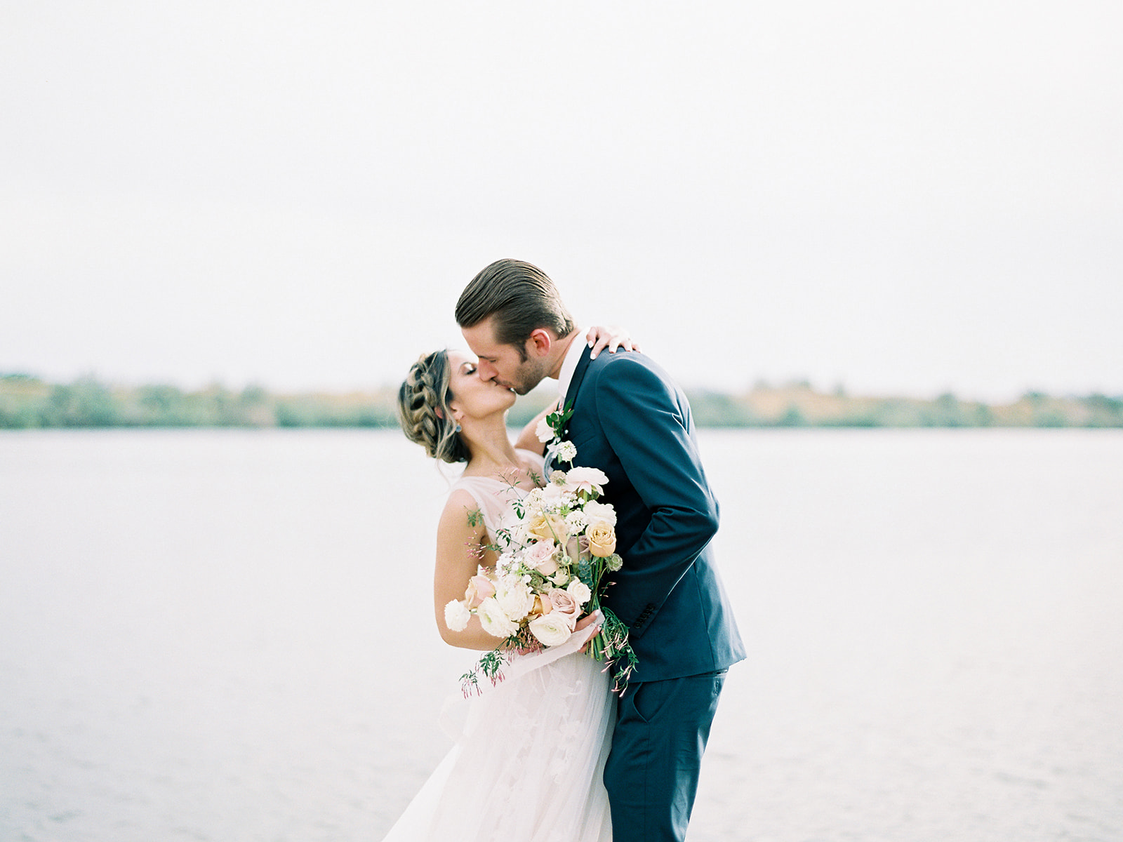 Bride and groom kissing with bride holding bouquet in front of lake.