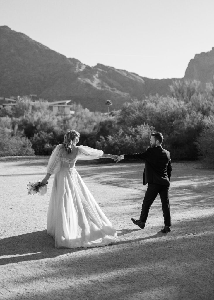Bride and groom walking holding hands with arms outstretched with desert mountains in the background.