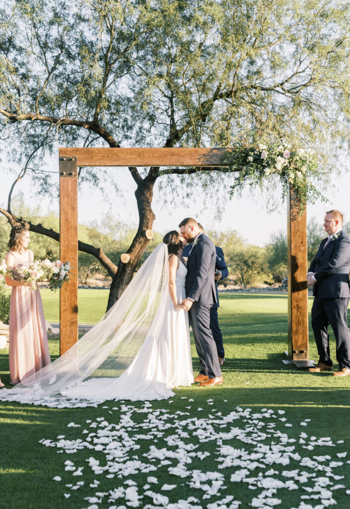 Bride and groom kissing under wood arch with floral and greenery attached in corner in outdoor ceremony space at McDowell Mountain.