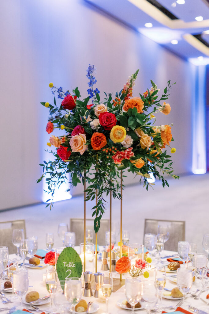 Tall wedding reception centerpiece on reception table of vibrant colors including fuchsia, yellow, peach, purple, and blue all on a gold stand.