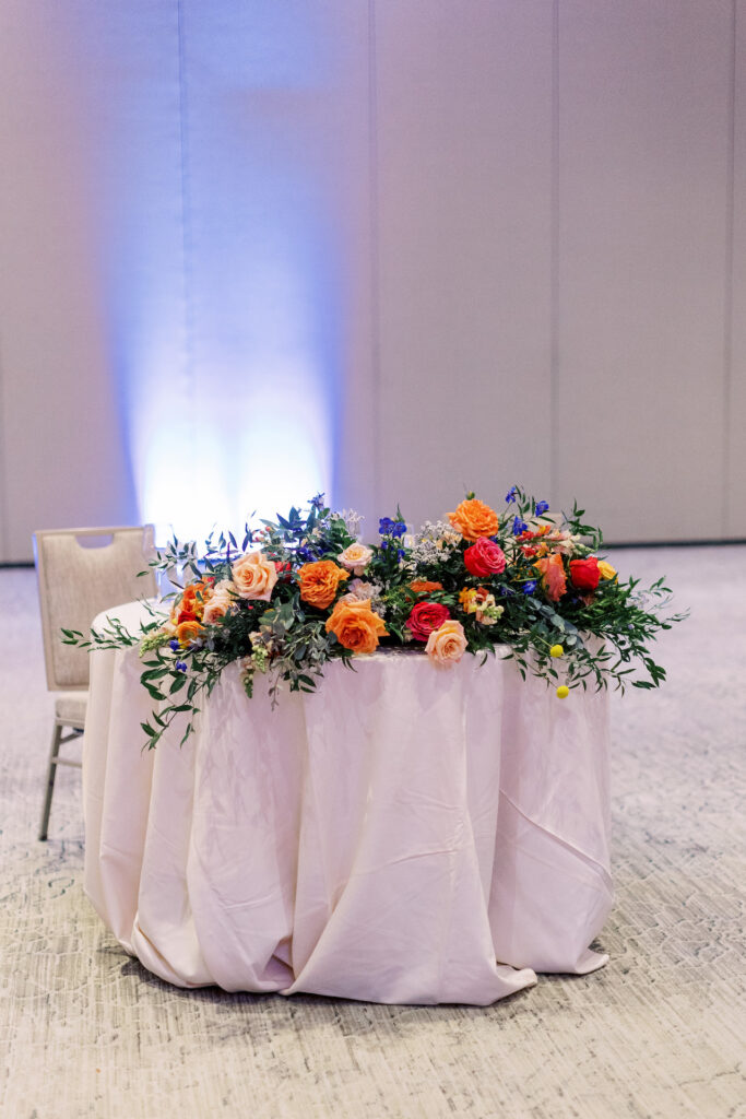 Indoor reception sweetheart table with a floral arrangement laying on front of table of vibrant colors including fuchsia, yellow, peach, purple, and blue.