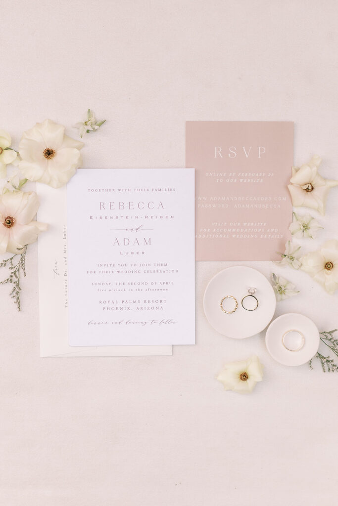Wedding invitation flat lay of white, cream and blush colors with white flowers and wedding rings.