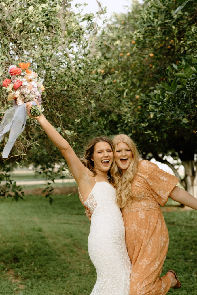 Bride and woman embracing, bride holding up bouquet, standing in grass field with trees.