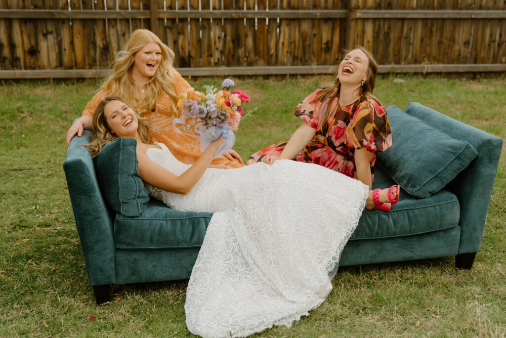 Bride sitting on teal couch in grass yard with two other women from Array Design, all laughing.