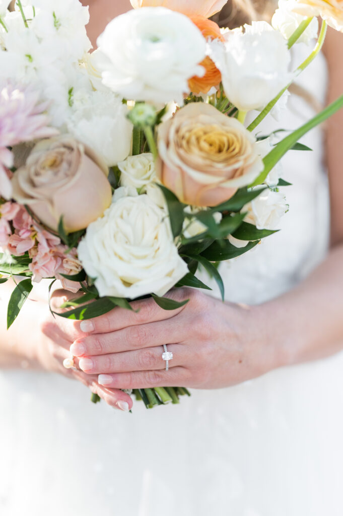 Bride's ring showcased with holding bouquet of white, pink, and yellow flowers.