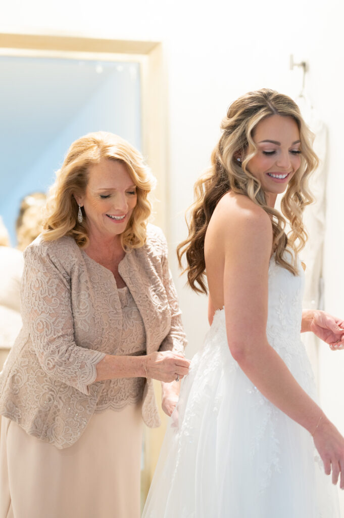 Woman in dark cream dress helping bride with zipping up back of dress, both smiling.