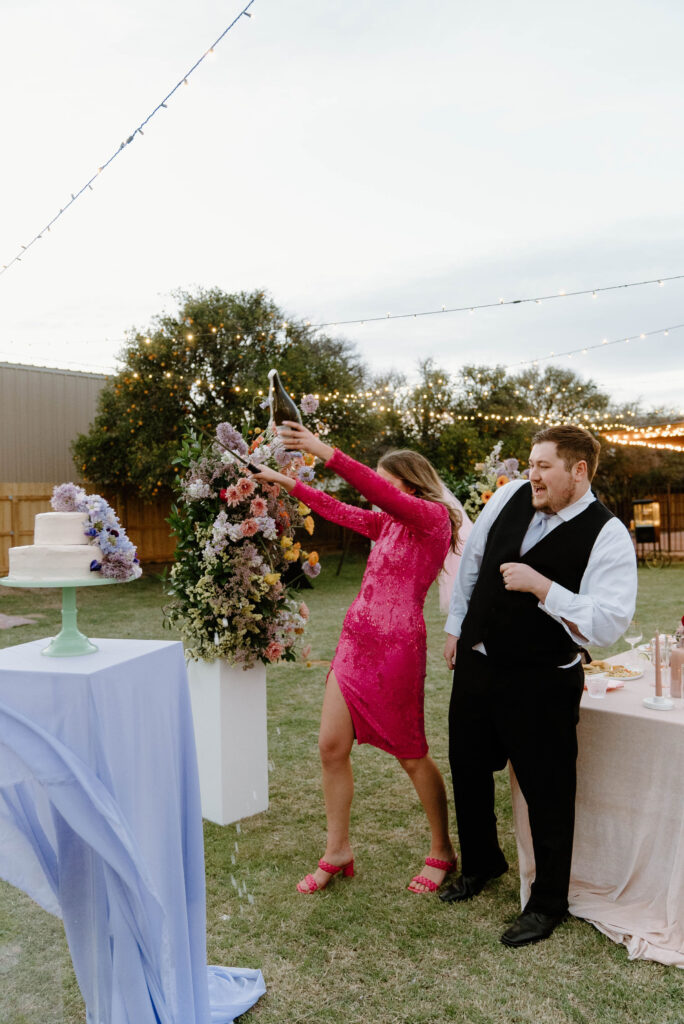 Bride opening champagne bottle with groom standing next to her.