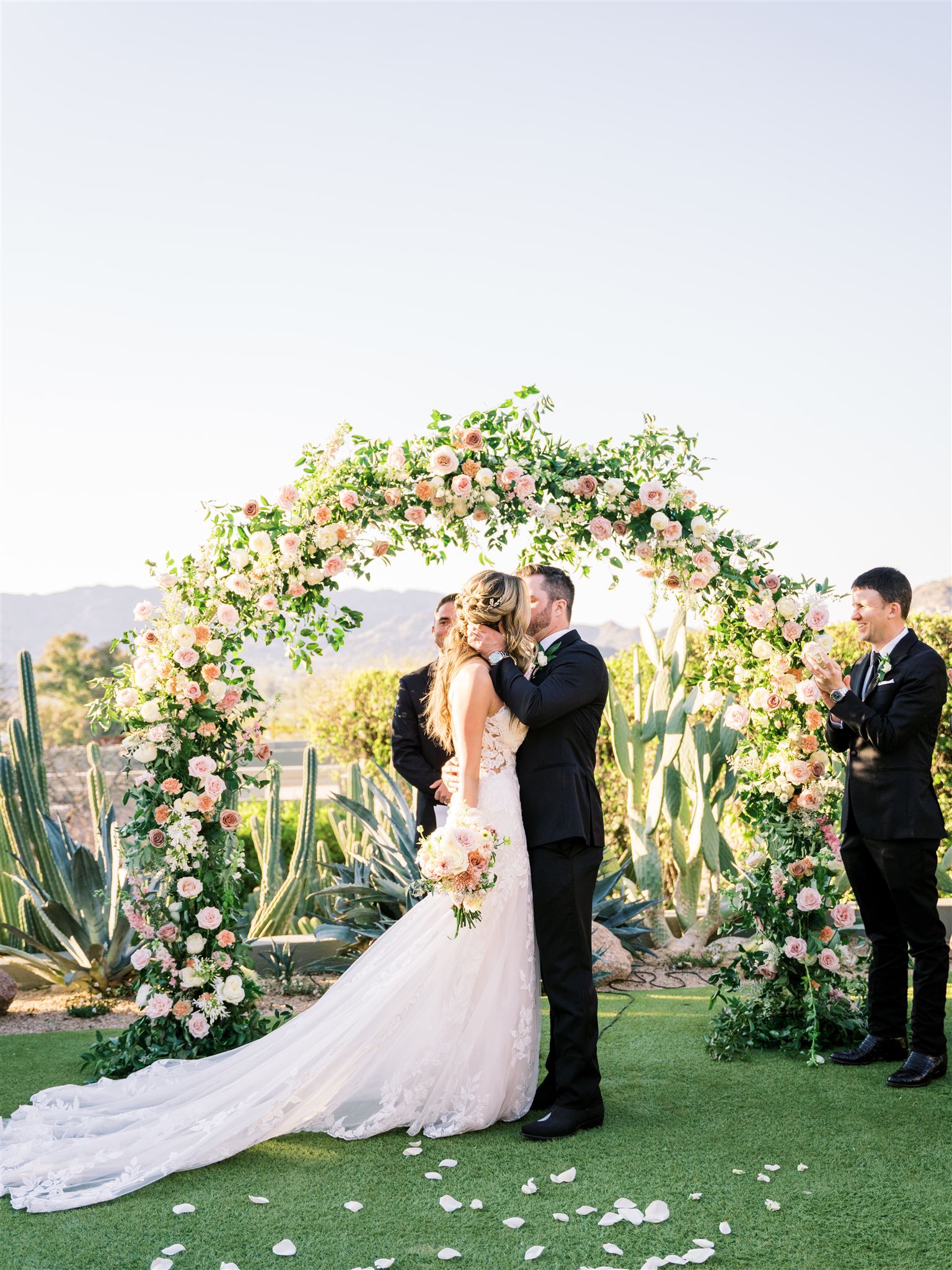 Bride and groom kissing under wedding ceremony arch of greenery and white and pink roses.