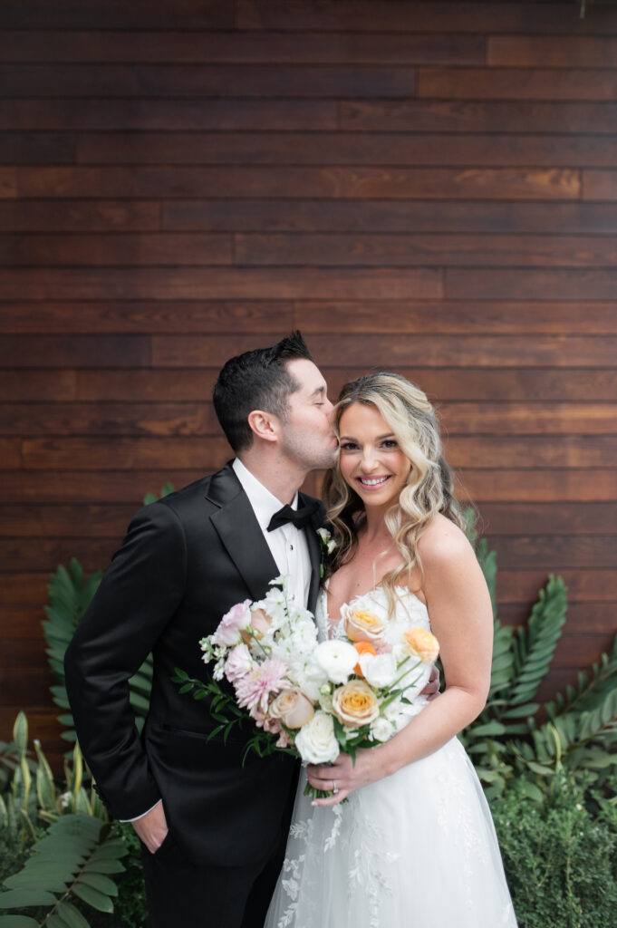 Groom kissing side of bride's face, while she is smiling, holding bridal bouquet in front of wood wall.