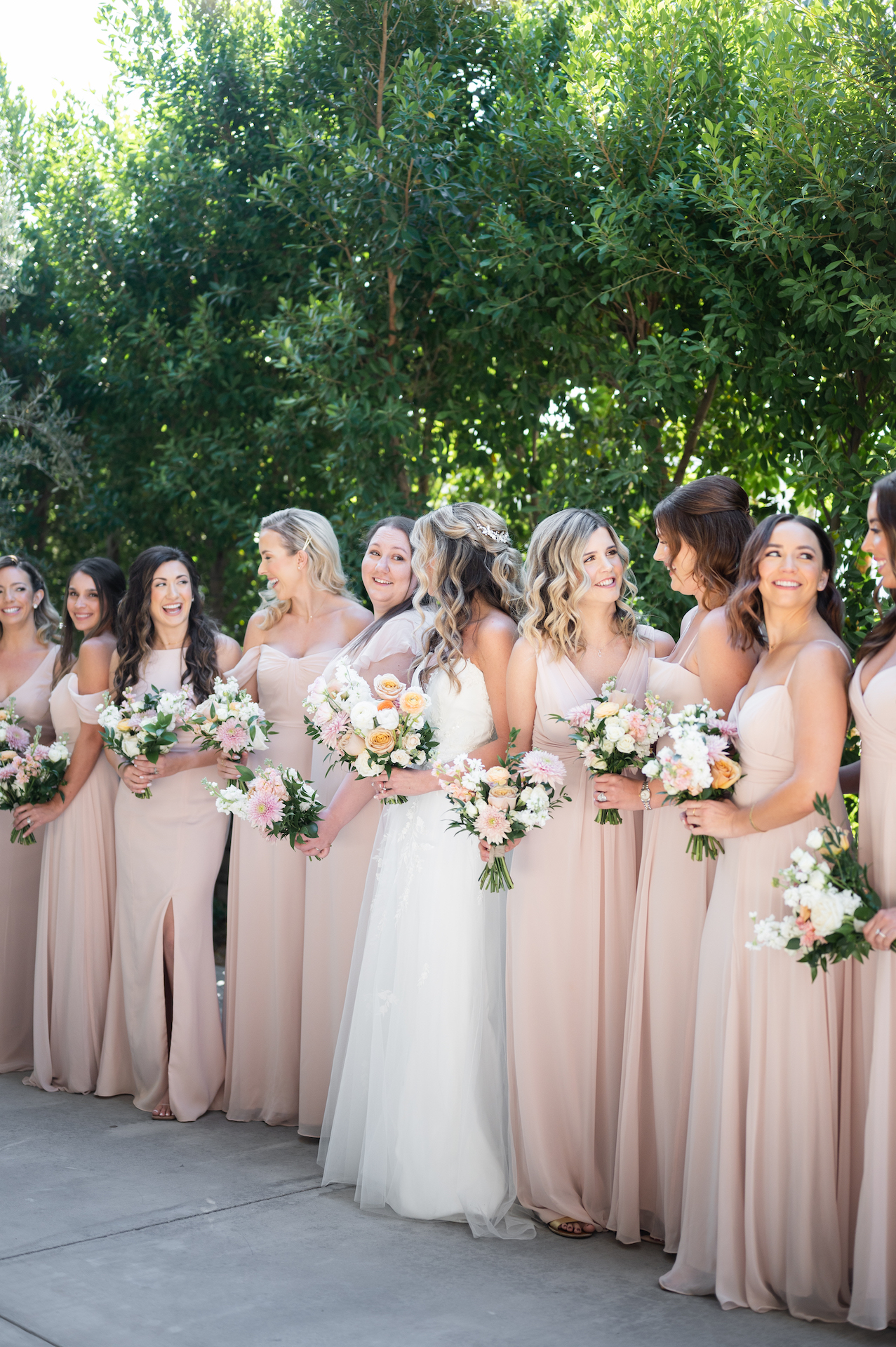 Bride and bridesmaids standing in row, smiling at each other. Bridesmaids in dusty pink dresses.